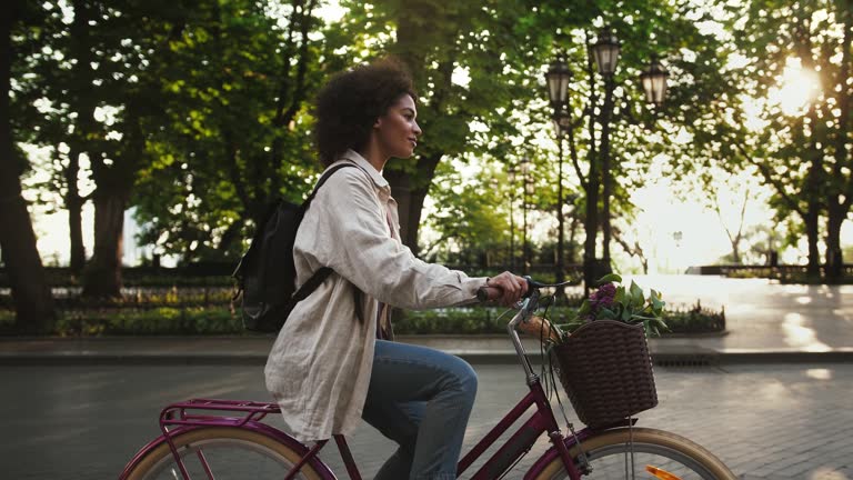 Afro-american woman riding a bicycle with flowers and baguette in basket by deserted city avenue next to an alley with green trees. Slow motion