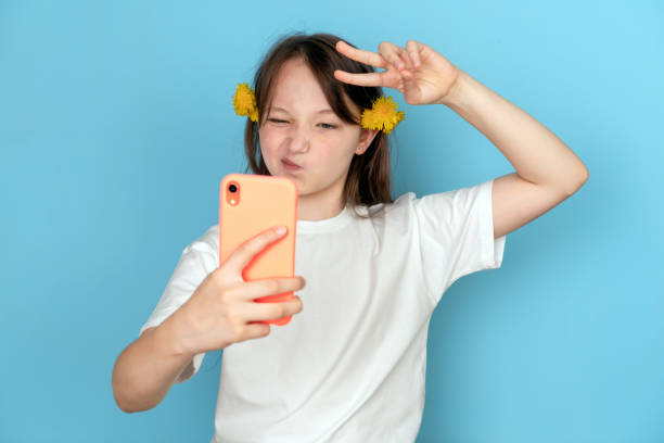 retrato de uma garota com dente-de-leão no cabelo e com um telefone nas mãos em um fundo azul. foto de estúdio. conceito de selfie e blogueiro. emoção de desprezo e o sinal de vitória - smiling humor child making a face - fotografias e filmes do acervo