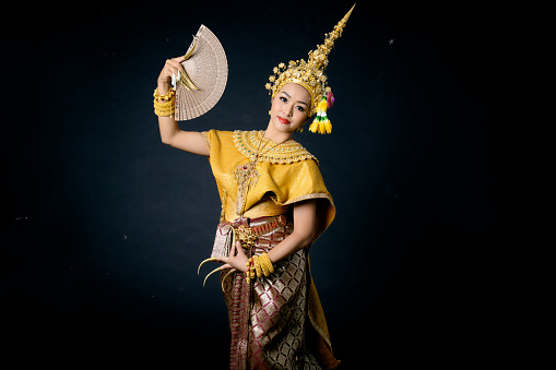 Beautiful Thai woman wearing traditional dancer dress headgear jewelry long nails holding fan dancing performance show on black background isolated.