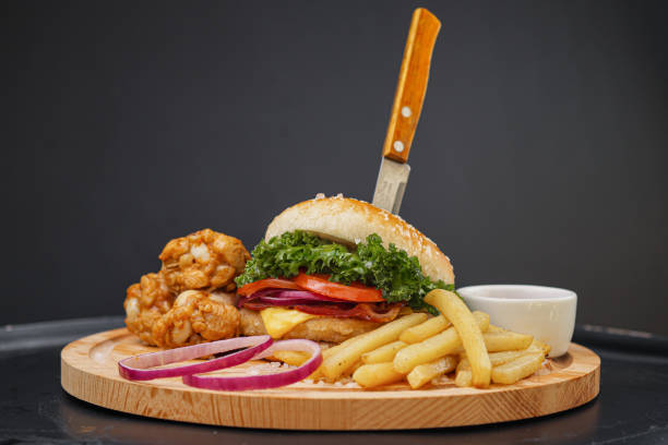 combo-set von fast food. saftiger hamburger, pommes frites, hühnerbeine und sauce. servierte holzbrett und besteck auf schwarzem hintergrund. fast-food-restaurant. lebensmittellieferung. - sandwich plate food french fries stock-fotos und bilder