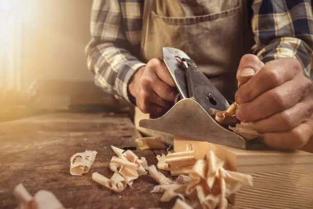 Close up of hands working with a manual metal planer on a rustic workbench. Chipped wood in the foreground.