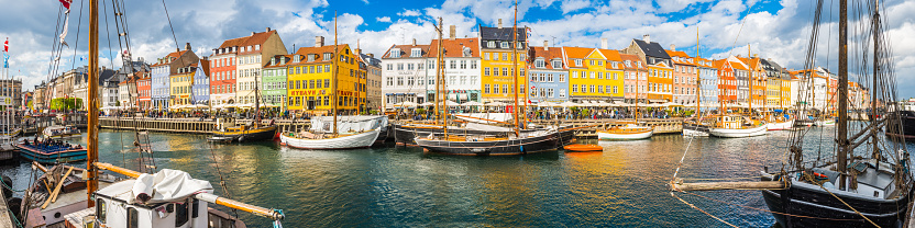 Crowds of people sitting, walking, chatting and relaxing on the picturesque waterfront of Nyhavn, its brightly coloured historic houses, cafes, bars, restaurants and nightclubs reflecting in the tranquil blue waters of the harbour canal leading to Kongens Nytor.