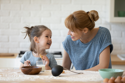 Smiling mother looking at little daughter and feeding her after bake cookies standing at table in kitchen. Happy caring mum and adorable girl eating flour and drink milk, preparing dinner.