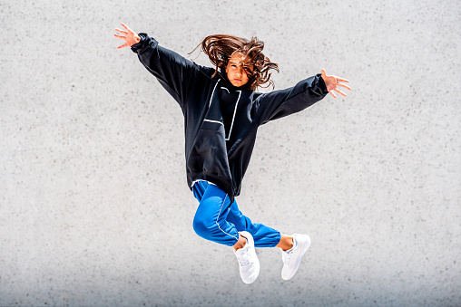 Young cute caucasian girl with long brown hair, wearing oversized black hoodie with white square on it, light blue sweat pants and white sneakers. She is posing in jump shot outdoors with white concrete wall in the background on a sunny day.