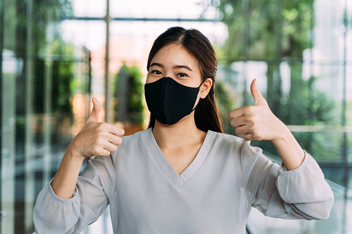 Joven estudiante asiática optimista dando gestos de pulgar hacia arriba. Lleva una máscara protectora (EPP) para evitar la contaminación del aire o la pandemia del virus Corona para mayor seguridad photo