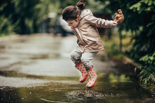 Smiling little girl playing in a puddle. Beautiful happy girl jumping in a puddle in a park