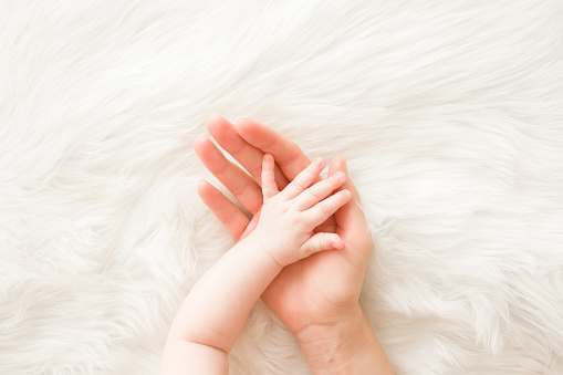 Infant hand on mother palm on white fluffy fur background. Lovely, emotional, sentimental moment. Trust and care concept. Closeup. Top down view.