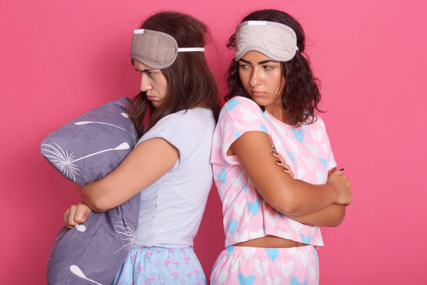 istock Two young charming girlfriends have fight during pajama party, posing back to back against rosy wall, dark haired women standing with pouty lips, being offended. 1227231172