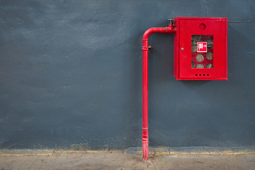 Fire hose in red box, pipe roll for fire emergency in red metal cabinet on gray painted concrete wall with tiled floor as part of firefighting system of industrial production plant, with copyspace.
