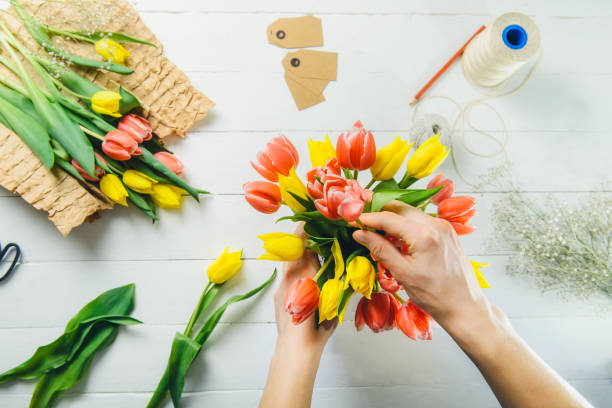Top view male florist hands making spring bouquet using tulips at workspace with recycled wrapping materials. Learning flower arranging. Education of floristry. Flowers delivery. Selective focus.