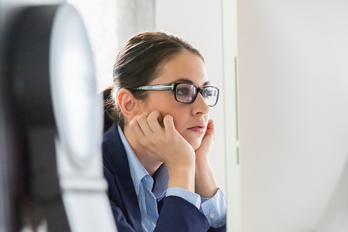 Boreuut. Young businesswoman sitting bored and sleepy with head in hands in front of her desktop computer screen reading e-mails and social media messages. Selective Focus. Natural Office Interior Light.