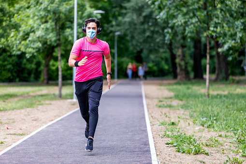 Sport during quarantine, self-isolation in the countryside. A young athletic guy is jogging on a dirt road in the meadow. He is wearing a blue medical mask and black  headphones