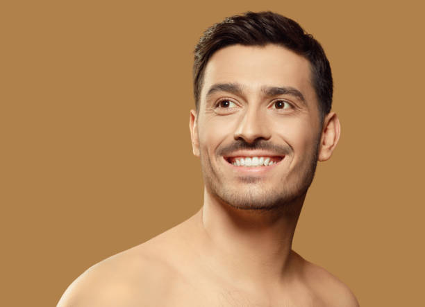 istock Young man isolated on light brown background, standing in front of camera, looking aside as if aspiring for good with happy smile on face. Men beauty skin care ads concept 1227242438