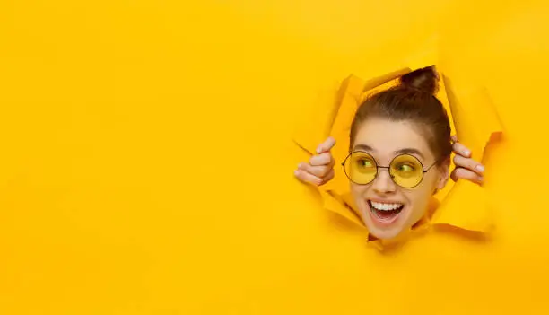 Photo of Horizontal banner of young girl in glasses tearing paper and peeking out hole, curious about commercial offer on copy space on left, isolated on yellow background
