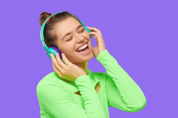 jovem feliz rindo menina ouvindo música em fones de ouvido sem fio com olhos fechados, vestido com top verde neon, se divertindo, isolado em fundo roxo - environmental conservation audio - fotografias e filmes do acervo