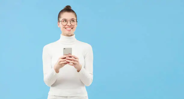 Photo of Horizontal banner of young student girl in glasses, looking aside with smile as if dreaming with smartphone in hands, isolated on blue background with copy space
