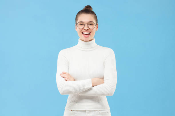 Young teenage girl dressed in white turtleneck, wearing glasses, standing with crossed arms and laughing happily, isolated on blue background Young teenage girl dressed in white turtleneck, wearing glasses, standing with crossed arms and laughing happily, isolated on blue background high collar stock pictures, royalty-free photos & images