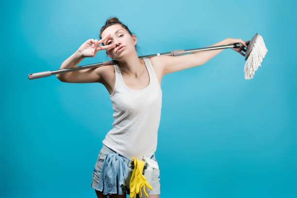 Housekeeper amusingly holds a mop around her neck like a rockstar microphone stand at a concert. Fun cleaning concept. Isolated on blue