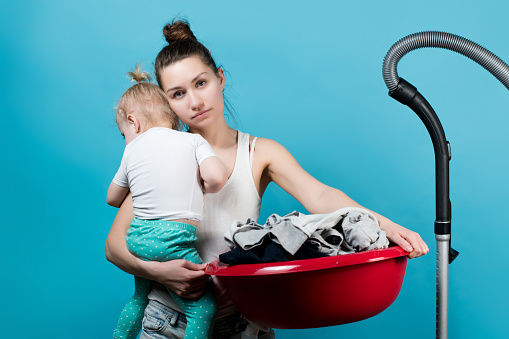 Mother wearily holds a small daughter and with her a large basin with clothes in the wash. Isolated on blue background.