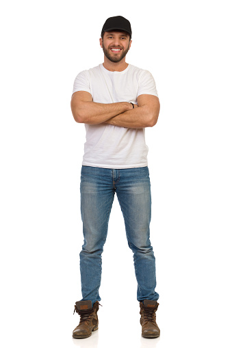 Confident smiling man in jeans, white t-shirt and black cap is standing with arms crossed. Full length studio shot isolated on white.