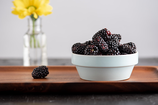 Bowl of fresh blackberries on wooden platter. Single blackberry on the side. Yellow flowers in vase in background. Gray background.