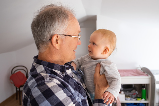 Grey-haired grandpa in eyeglasses holding cute baby girl. Grandfather and grandchild looking at each other. Help from grandparents and staying at home concept