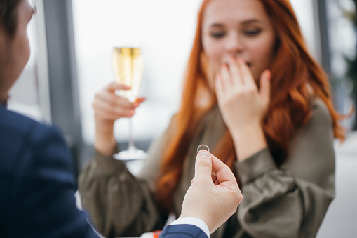 romantic date of lovely caucasian couple in rich restaurant, redhaired lady stay in shock after marriage proposal by her boyfrined in tuxedo, he hold golden ring in hands. love, couple, marriage, anniversary concept