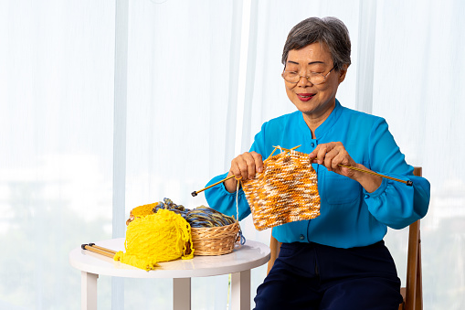 Asian mature senior woman knitting wool clothes and sitting on wooden chair, elderly generation hobby activity