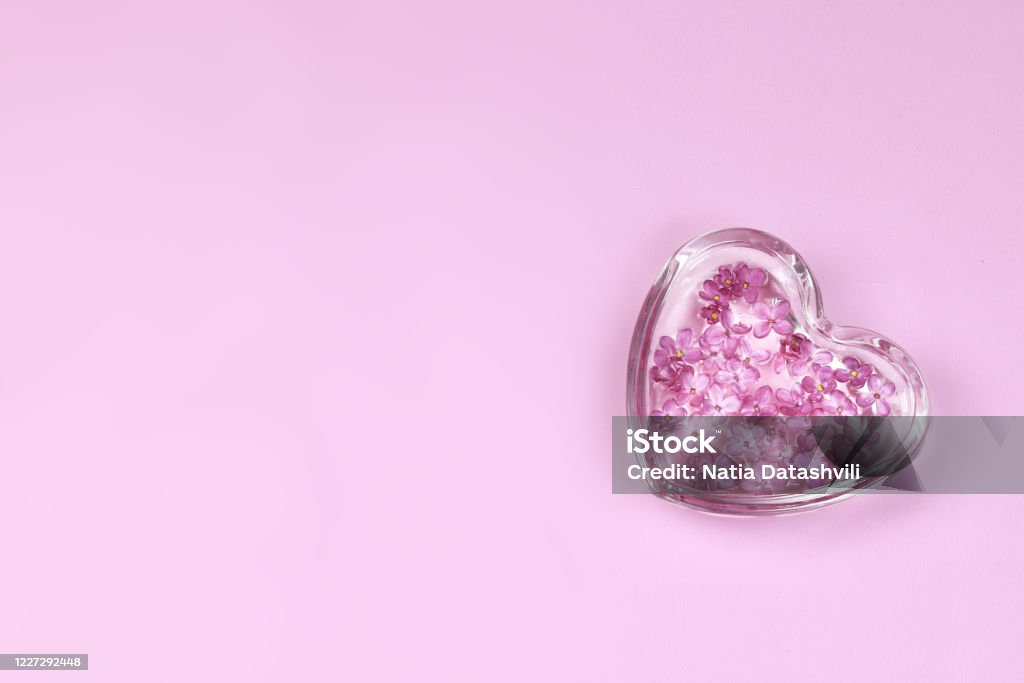 Spa concept plat lay image. Heart shaped glass plate with water and lilac flowers on light pink background. Spa concept plat lay image. Heart Shape Stock Photo