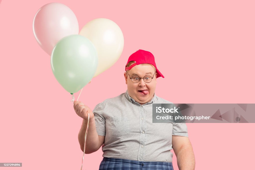 Funny man much similar to Winnie the Pooh with air baloons isolated over pink Portrait of plump children animator with comic crazy face expression wearing red baseball cap, holding colorful air balloons, looks at camera like a ninny. People, celebration concept Adult Stock Photo