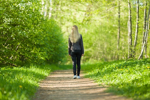 One young blonde woman slowly walking on natural trail in green forest. Warm sunny spring day. Spending time alone in nature. Peaceful atmosphere. Back view.