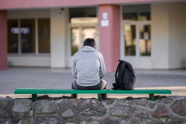 un joven sentado en el banco del patio de la escuela. tiempo de descanso. vista trasera. - solitario fotografías e imágenes de stock
