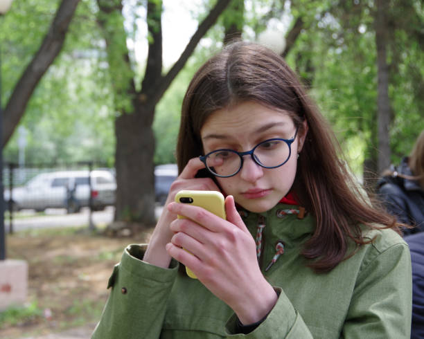 front view of a sad cute caucasian girl in blue glasses and a green jacket wiping a tear when looking at the phone - tear beautiful despair anxiety imagens e fotografias de stock