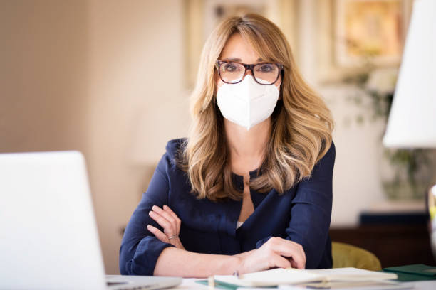 Middle aged woman wearing face mask for prevention while working from home Portrait shot of middle aged woman wearing face mask while working at home on her notebook and text messaging during coronavirus pandemic. secretary photos stock pictures, royalty-free photos & images
