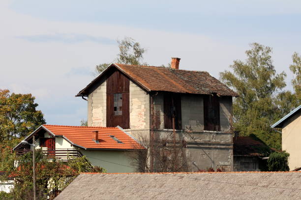 Abandoned strange looking old building with destroyed roof with leaning chimney and broken windows next to small attached newly built suburban family house surrounded with trees and other houses