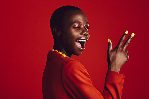 Excited female model winking an eye with finger gun on red background. African female model with buzz cut hairstyle.