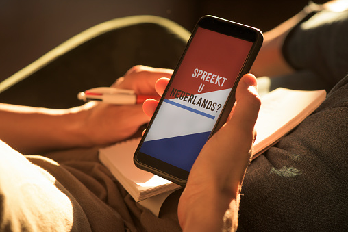 closeup of a young caucasian man, wearing casual clothes, sitting on an armchair, having his smartphone in his hands with the text do you speak Dutch and the flag of the Netherlands in its screen