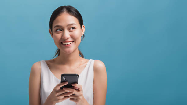 asiática feliz hermosas mujeres blogger sonriendo y sosteniendo el teléfono inteligente aislado en fondo de color azul con espacio de copia. concepto de marketing tecnológico online. - teenage girls cheerful smiling one person fotografías e imágenes de stock