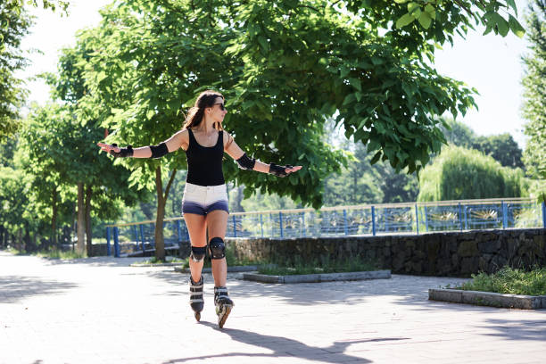 Roller skating in park Young pretty brunette woman, riding rollerblades in city park with green trees. Fit sporty girl, wearing protective outfit, roller-skating in summer. Full-length portrait of slim sportswoman. inline skating stock pictures, royalty-free photos & images