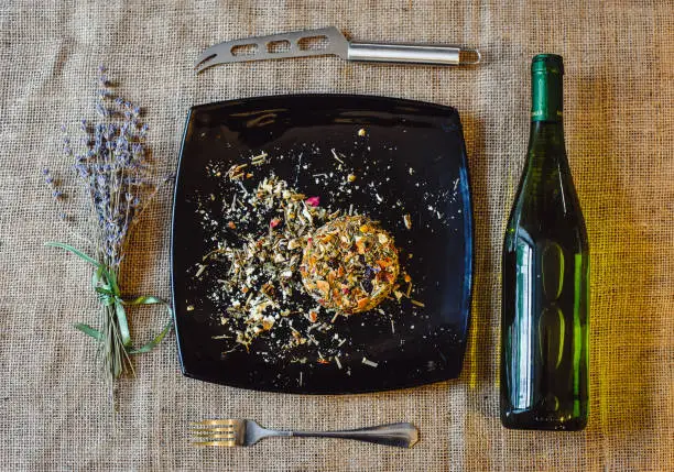 The round head of the French spicy cheese Boulette d'Avesnes sprinkled with different herbs, lies on a black plate. Arrangement: boulette d'avesnes cheese, wine bottle, knife, lavender bouquet