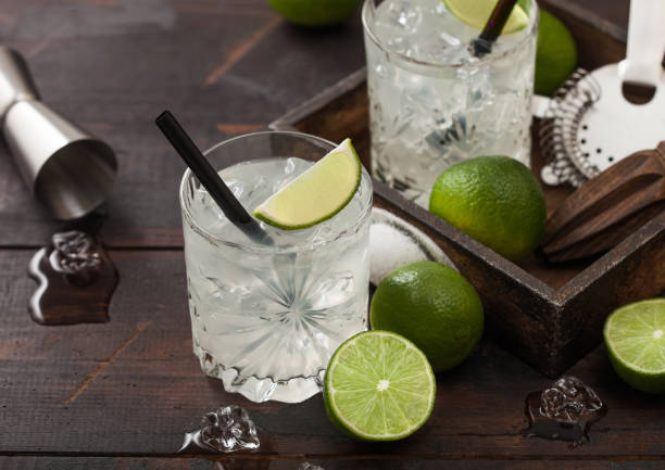 Gimlet Kamikaze cocktail in crystal glasses with lime slice and ice on wooden board with fresh limes and jigger with strainer. stock photo