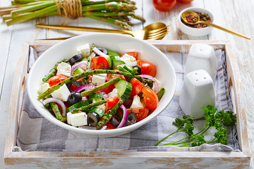 Greek salad of asparagus, feta cheese, chopped tomatoes, cucumbers, black olives, red onion, served on a white bowl on a serving tray with a bunch of asparagus spears, parsley, peppercorns, close-up