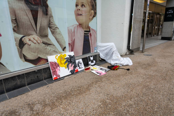 A homeless artist trying to sell some of their art works to make money. There is a contrasting image in the store window behind the art. Oxford, Oxfordshire, England, UK.
