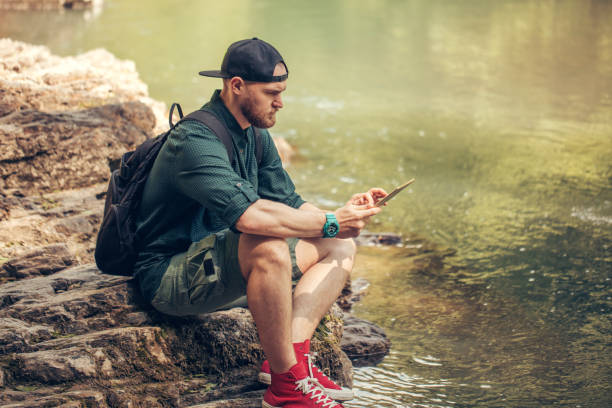 istock Single male hiker using tablet on nature while sitting on rocky river shore 1227347218
