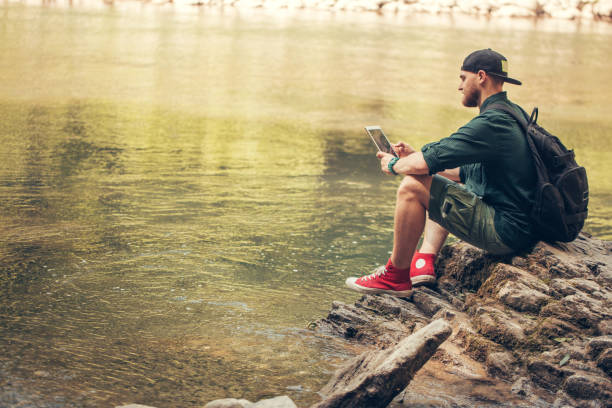 istock Single male hiker using tablet on nature while sitting on rocky river shore 1227347494
