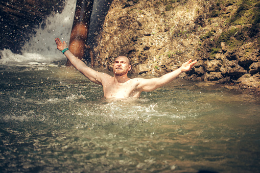A caucasian bearded tourist bathing in the rocky mountain lake, refreshing himself after a long hike in summer highland forests in Nature Reserve.