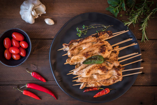 Plate with grilled chicken skewers, garlic, tomatoes, herbs and red chili on dark wood. Top view