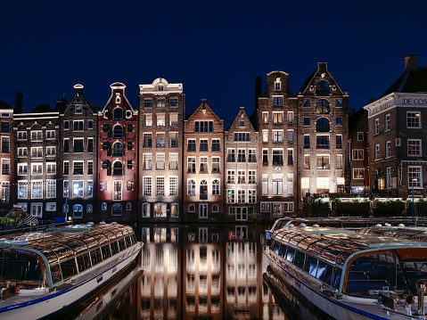 Set of neighbouring buildings downtown Amsterdam, in front of a canal - Damrak. Night photograph with sightseeing boats.
