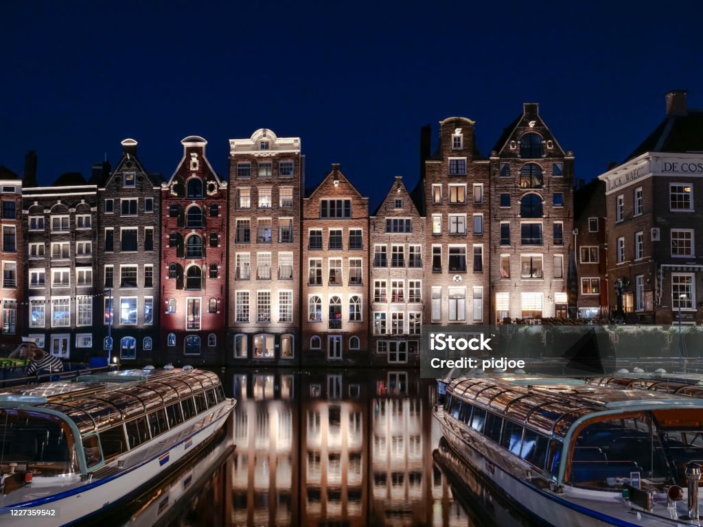 Amsterdam skyline old buildings waterfront Damrak Set of neighbouring buildings downtown Amsterdam, in front of a canal - Damrak. Night photograph with sightseeing boats. Amsterdam Stock Photo
