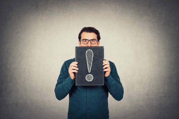 Shocked businessman looking astonished with big eyes, holding a board with a drawn exclamation mark isolated on grey wall background. Business worker wearing glasses, has doubts, panic concept. un hombre de negocios sorprendido que se asombraba de grandes ojos, sosteniendo un tablero con un signo de exclamación dibujado aislado en el fondo gris de la pared. un trabajador de negocios que lleva gafas, tiene dudas, concepto de pánico.